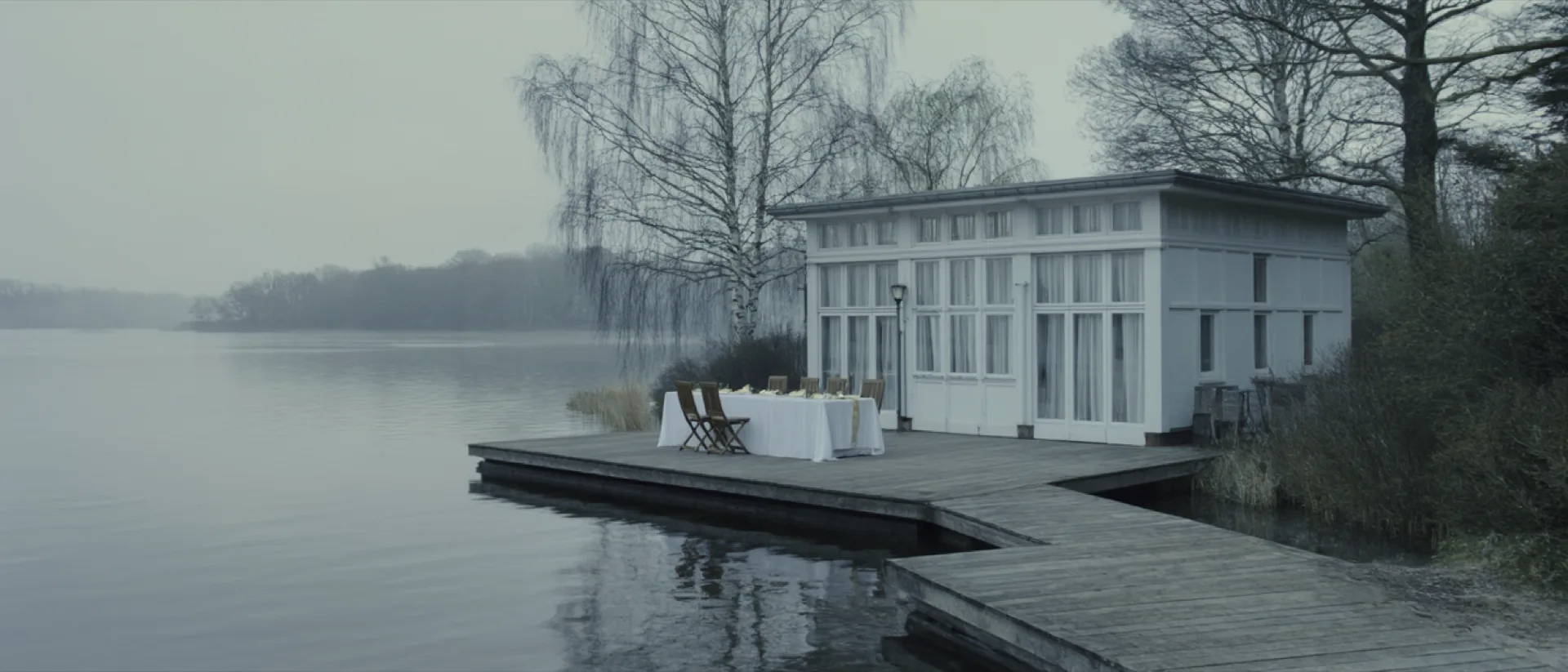The Trauwerk boathouse at Weißensee on an overcast morning, a long linen-dressed table on the timber terrace above still water